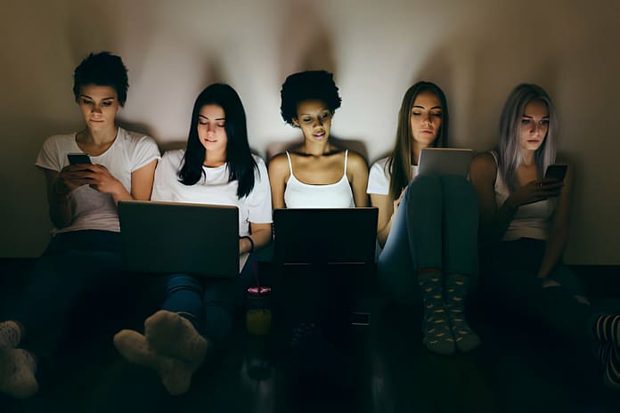 Group of female friends at home sitting on floor using mobile phone and laptops in the dark